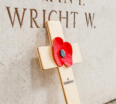 A small wooden cross has a poppy in the middle and is leaning against a Rememberance Day wall of names of people who died in the war.